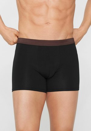 Männlicher Torso trägt schwarze Boxershorts mit braunem Bund, Hände richten den Bund auf beiden Seiten aus.