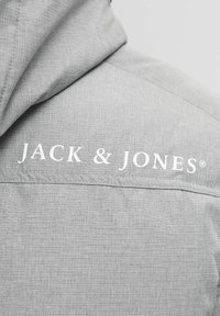 Grå hættejakke med struktureret stof og et hvidt "JACK & JONES" logo trykt på ryggen nær halsudskæringen.