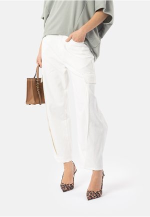 Persona che indossa pantaloni bianchi a gamba larga, top grigio largo, tacchi alti con stampa leopardata e tiene una borsa marrone con una mano in tasca.