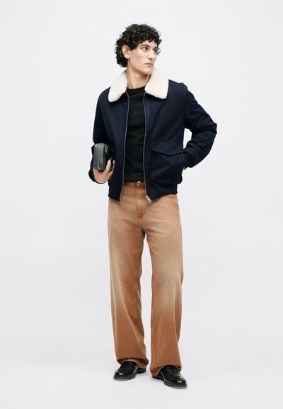 Temno modra bomber jakna s kremno umetno krznenim ovratnikom, črna majica, bež široke hlače in črni čevlji, drži majhno črno torbico.