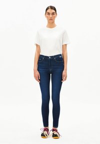 Dunkelblaue Skinny-Jeans aus Denim, hoch tailliert, mit Vorderentaschen und einem Knopfverschluss, kombiniert mit einem schlichten weißen T-Shirt und schwarzen Schuhen.