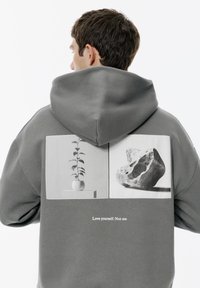 Mężczyzna w szarej bluzie z kapturem, pokazujący plecy, z czarno-białymi zdjęciami doniczkowej rośliny i kamienia, tekst brzmi: "Kochaj siebie. Nie mnie."