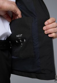 Chaqueta negra con un acabado suave y mate, que presenta un bolsillo con una etiqueta que muestra "NEXT" y símbolos de cuidado. Detalle de cremallera visible en el dobladillo.