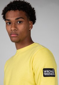 Sudadera de algodón amarilla con cuello redondo y puños acanalados, que presenta un parche negro con el texto "WRONG FRIENDS" en la manga izquierda.