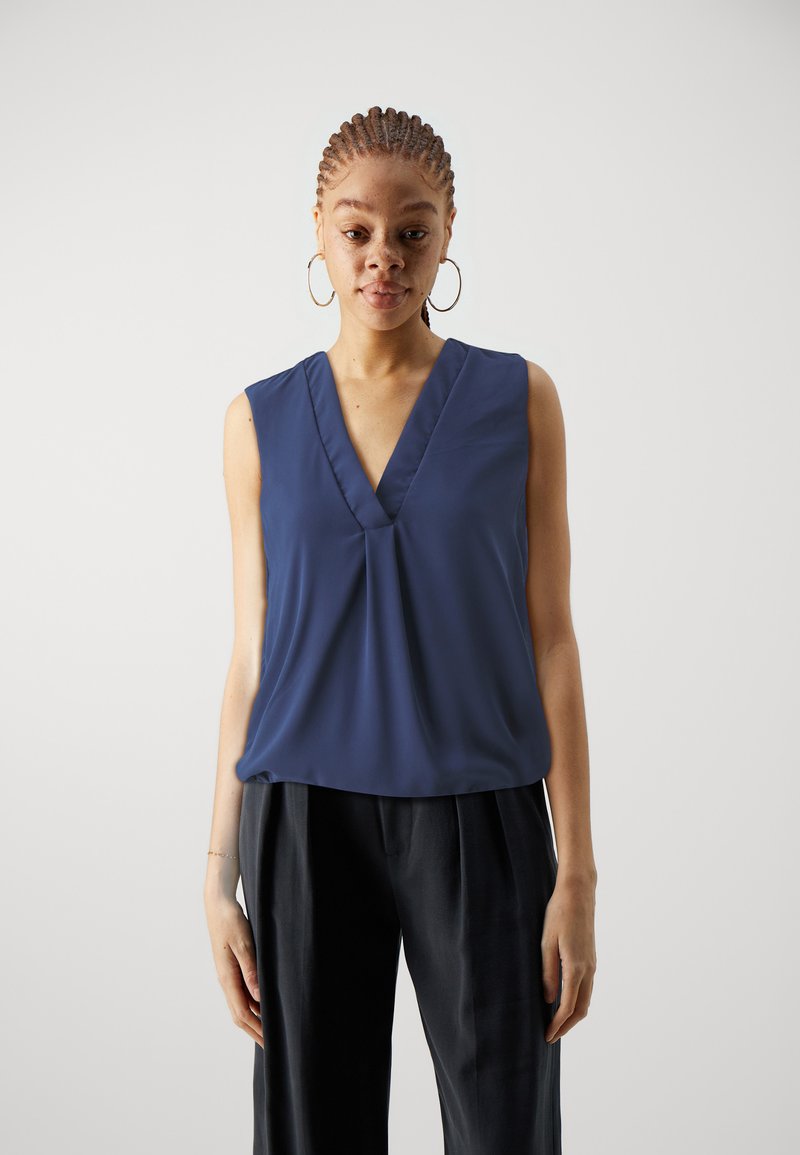 Top senza maniche di colore blu navy con scollatura a V, caratterizzato da tessuto drappeggiato e una vestibilità comoda. Abbinato a pantaloni scuri e larghi.