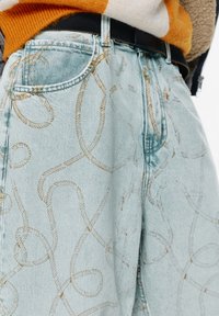 Jeans din denim de culoare albastru deschis cu un model de frânghie răsucită din fir de aur. Caracteristici includ buzunare laterale și o curea neagră. Design texturat, casual.