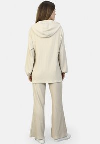 Beige Kapuzenoberteil aus strukturiertem Stoff mit langen Ärmeln, kombiniert mit passender, weit geschnittenen Hose. Die Schuhe sind hellfarbene Sneakers.