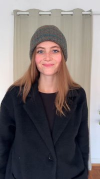 Čierny, textúrovaný kabát s jedným gombíkom, nosený cez čiernu košeľu. Hnedá a tyrkysová čiapka. Svetlá drevená podlaha, neutrálne pozadie.