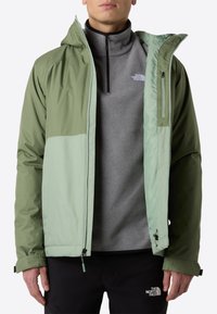 Vihreä takki, jossa on vaaleamman vihreät yksityiskohdat, valmistettu sileästä kankaasta. Takissa on vetskarilla varustettu etuosa, huppu ja säädettävät hihansuut, jotka ovat harmaan fleecetakin päällä.