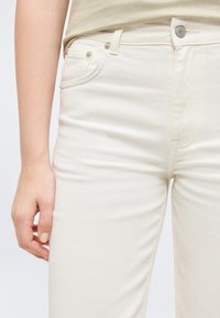 Κοντινό πλάνο ενός ατόμου που φοράει τζιν υψηλής μέσης σε εκρού χρώμα και μπεζ τοπ, με το αριστερό χέρι χαλαρό δίπλα στο σώμα.