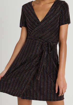 Kobieta w krótkim rękawie, kopertowej sukience z dekoltem w serek, wielokolorowymi pionowymi paskami i paskiem do wiązania, trzyma delikatnie uniesioną spódnicę.