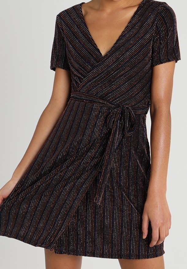 Femme portant une robe portefeuille manches courtes à col en V avec des rayures verticales multicolores et une ceinture à nouer, tenant la jupe légèrement relevée.