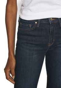 Persona que lleva jeans de mezclilla azul oscuro y una camisa blanca metida por dentro, con un brazo relajado apoyado a su lado.