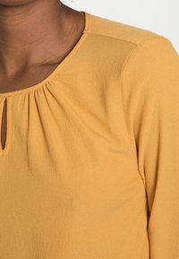Blusa gialla con scollo a barca, dettaglio plissettato sul petto, texture morbida e silhouette aderente. Nessuna cucitura o decorazione visibile.