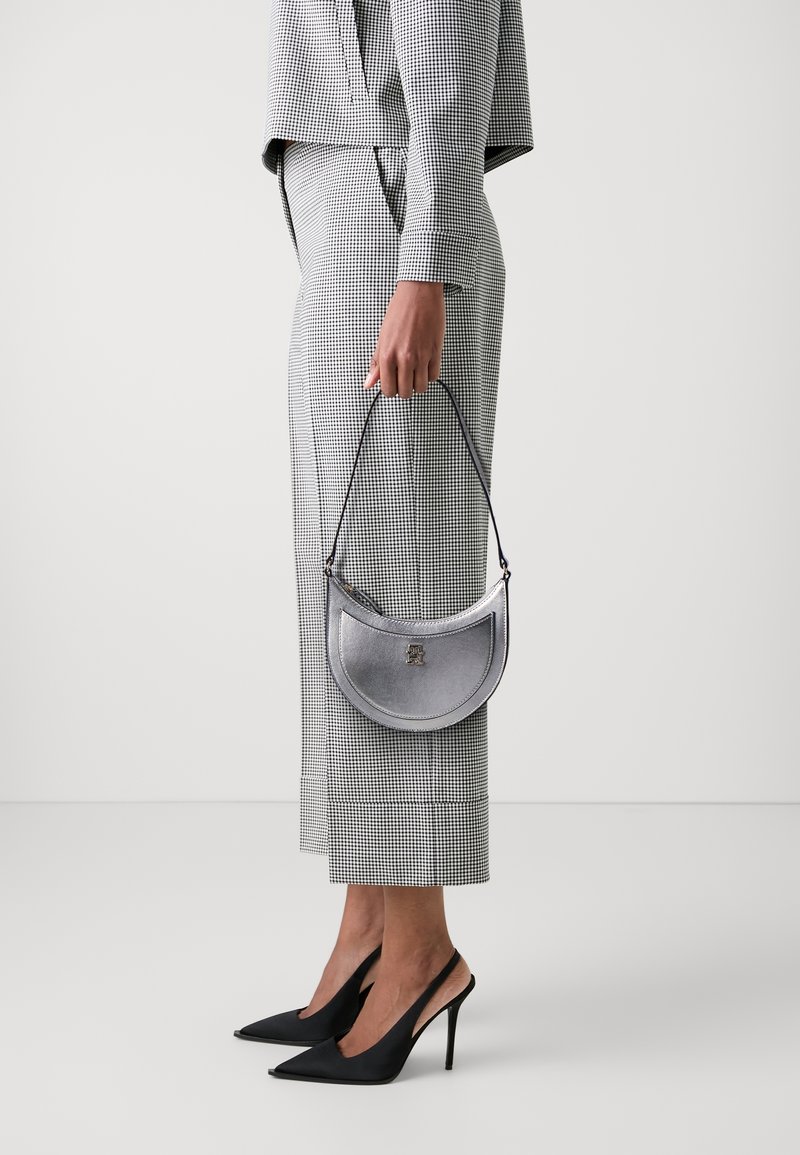 Personne portant un tailleur-pantalon à carreaux noirs et blancs, tenant un sac à main argenté en forme de croissant, portant des chaussures slingback à talons aiguilles pointues noires.