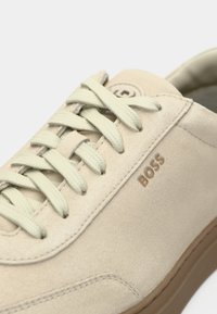 Beežid seemisnahast vabaajajalatsid heleroheliste paeltega, külje peal reljeefne logo, tekstureeritud pealne ja kummist tald. Lähedalt vaadatuna, mis rõhutab detaile.