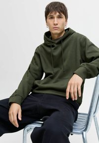 Olive green hoodie with a kangaroo pocket and drawstrings, paired with dark pants. Model sits on a metal chair against a neutral background.