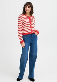Cardigan a righe rosse e bianche con bottoni, polsini e orlo a coste. Indossato sopra jeans a gamba larga blu. Morbido texture a maglia.