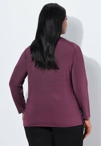 Frau mit langen schwarzen Haaren, die ein lila Oberteil mit langen Ärmeln und schwarze Hosen trägt, mit dem Rücken zu einer schlichten, hellen Hintergrund. 