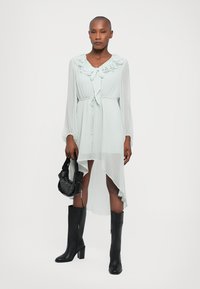 Rochie lungă de culoare verde deschis, cu mâneci lungi, guler plisat, talie ajustabilă cu șnur, hem asymetric și material transparent; asortată cu cizma negre până la genunchi și o geantă mică neagră.