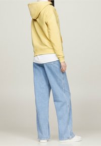 Felpa gialla con vestibilità ampia e cappuccio regolabile, abbinata a jeans a gamba larga blu chiaro e sneakers bianche. Tessuto morbido.