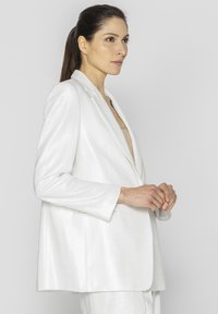 Biała marynarka wykonana z błyszczącego materiału, o luźnym kroju, z wyprofilowanym kołnierzem i długimi rękawami, z minimalnymi detalami i bez zapięcia.