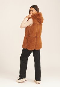 Faux fur vest in rich orange-brown, featuring a hood, sleeveless design, and a soft texture. Paired with black pants and sneakers.