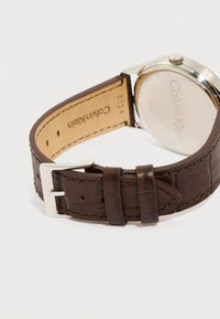 Καφέ δερμάτινο λουράκι ρολογιού Calvin Klein με ασημένια αγκράφα και λεπτομέρειες ραφών, μερικώς ανοιχτό που δείχνει το χαραγμένο μεταλλικό πίσω μέρος του ρολογιού.