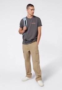 Camiseta gris con mangas enrolladas y logo naranja, combinada con pantalones cargo beige con bolsillos. Accesorios incluyen una mochila azul.