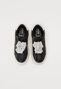 Svarte lær sneakers med hvit gummisåle, og tre hvite tekstilblomster på tungen, samt merkevaren på sålen.