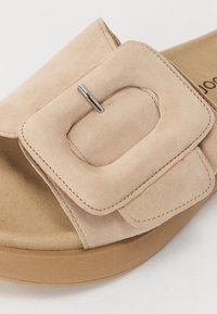 Beige suede slide sandal featuring a prominent square buckle with a silver accent. Cushioned footbed and flat, light brown sole.