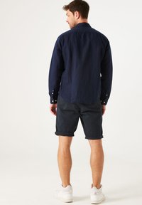 Camisa de manga larga azul marino de tela texturizada, combinada con pantalones cortos oscuros arremangados y zapatillas blancas, vista desde la parte trasera.