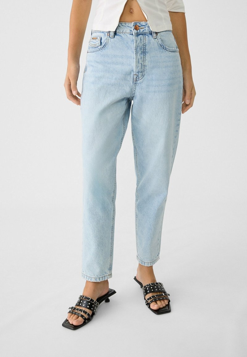 Jeans de tiro alto en azul claro con un corte relajado, que cuentan con bolsillos frontales, cierre de cremallera y un sutil desgaste. Combinados con sandalias de tacón negro.