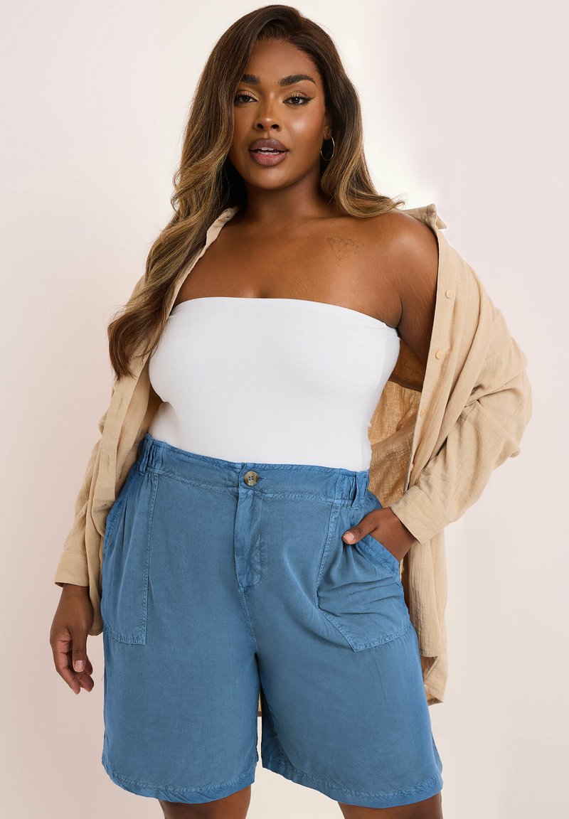 Ljust blå shorts i mjukt tyg, med knappstängning och sidofickor. Bärs med en vit axelbandslös topp och en knäppt beige skjorta.