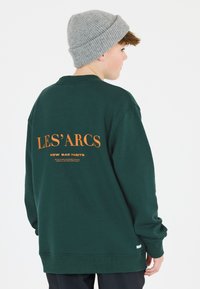 Grön sweatshirt med ribbade muddar, med orange text på ryggen: "LES' ARCS NEW BAD HABITS." Åtföljs av en grå stickad mössa.