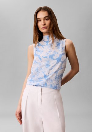 Kobieta z długimi brązowymi włosami, ubrana w bezrękawnikowy niebiesko-biały wzorzysty top i białe spodnie z wysokim stanem, stojąca na szarym tle.