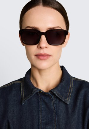 Femme aux cheveux bruns tirés en arrière porte de grandes lunettes de soleil noires carrées et une veste en jean foncé avec des coutures jaunes au col.