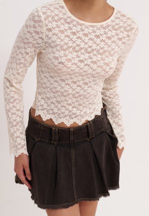 Femme portant un haut en dentelle blanche à manches longues avec des bords festonnés et une mini-jupe en jean noire plissée avec un ourlet effiloché.