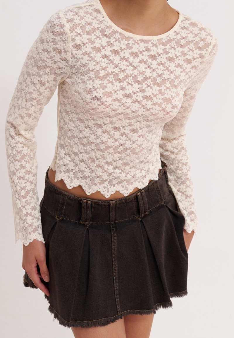 Femme portant un haut en dentelle blanche à manches longues avec des bords festonnés et une mini-jupe en jean noire plissée avec un ourlet effiloché.