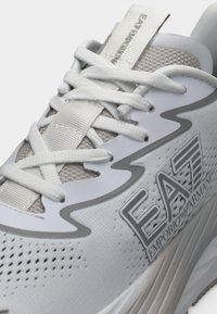 Λευκό και γκρι sneaker Emporio Armani EA7 με επάνω μέρος από δίχτυ, λεπτομερείς κορδόνια και υφασμένο πέλμα, κοντινό πλάνο.
