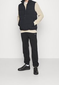 Černá péřová vesta přes smetanový pletený svetr, doplněná černými teplákovými kalhotami a černými teniskami. Bílé detaily na ponožkách. Ležérní outfit.