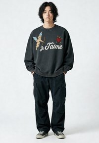 Jeune homme debout les mains dans les poches, portant un pantalon cargo noir, des baskets noires et un sweat-shirt foncé avec un cupidon et le texte "Je T'aime".