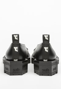 Scarpe in pelle nera con una suola nera spessa e testurizzata. Presentano linguette di marca con un motivo del logo sul retro. Design moderno e minimalista.