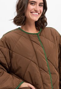 Smeđe prešano vanjsko odijelo s dijamantnim uzorkom i zelenim obrubom. Ima okrugli izrez i široke rukave. Mekana tekstura, srednje dugo fit.