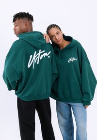Temno zeleni oversize hoodiji s sprednjim žepom, belim logotipom na prsih in hrbtu, noseni z črnimi in svetlo modrimi kavbojkami.