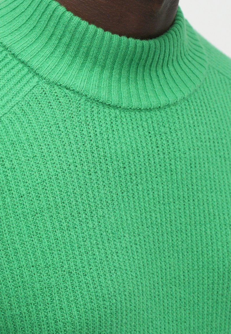 Pull en tricot vert avec une texture côtelée et un col haut. Présente une coupe près du corps avec des mailles uniformes sur l'ensemble.