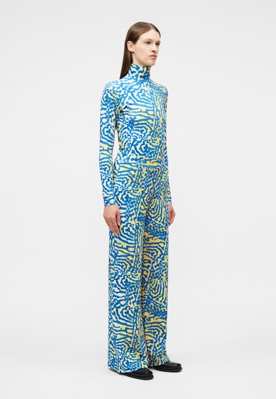 Γυναίκα όρθια που φοράει μπλούζα με υψηλό γιακά σε μπλε και κίτρινο με αφηρημένο μοτίβο και παντελόνι καμπάνα που ταιριάζει, με μαύρα παπούτσια.