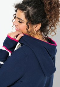 Granatowy sweter z kapturem, wykończony różowym kapturem, z białymi i różowymi paskami na rękawach. Teksturowany materiał, luźny krój.