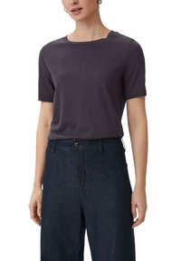 Kobieta w ciemnofioletowej krótkiej dzianinowej bluzce włożonej w wysokie, ciemnoniebieskie, szerokie spodnie dżinsowe na białym tle.