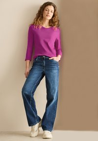 Fuchsia lange mouwtop met een ronde hals, gecombineerd met losse pasvorm blauwe denim jeans en beige sneakers, tegen een neutrale achtergrond.